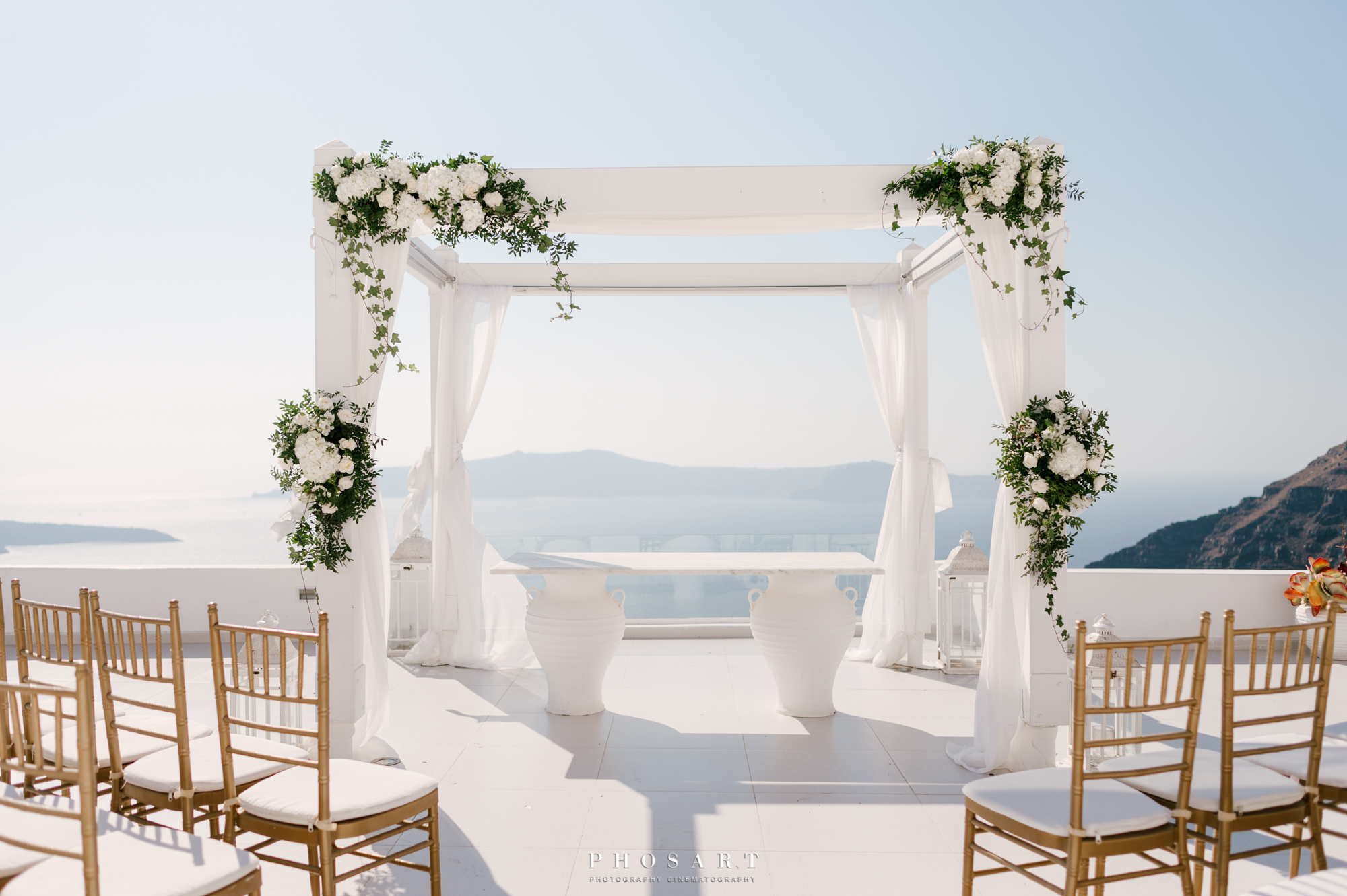A white wooden wedding gazebo overlooking the Aegean, decorated with white flowers and wooden gold empty chairs