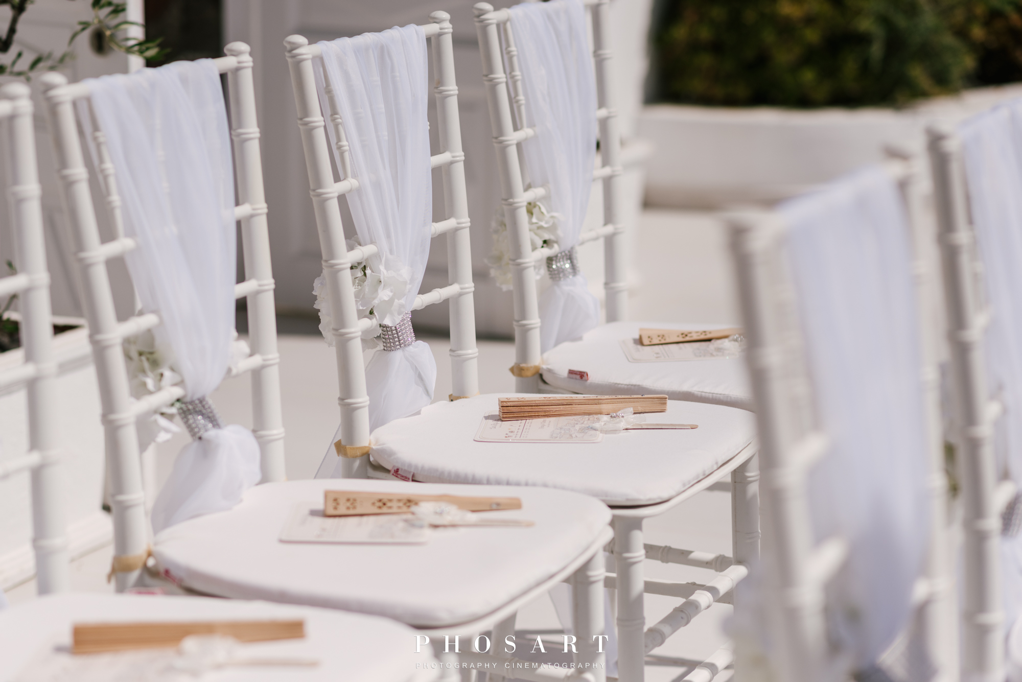 three white wooden chairs ornated with white fabrics, flowers and elegant hand fans