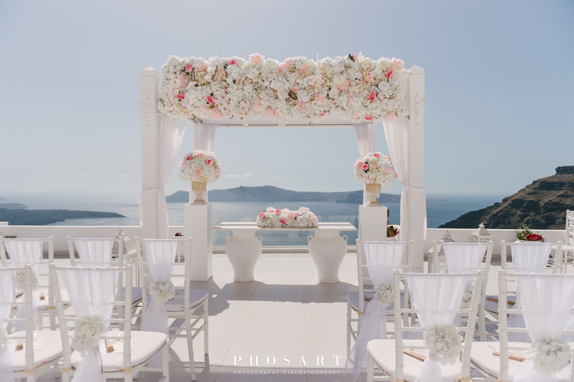 a white wooden gazebo overlooking the Aegean, decorated with white & pink flowers and wooden white empty chairs before the wedding ceremony