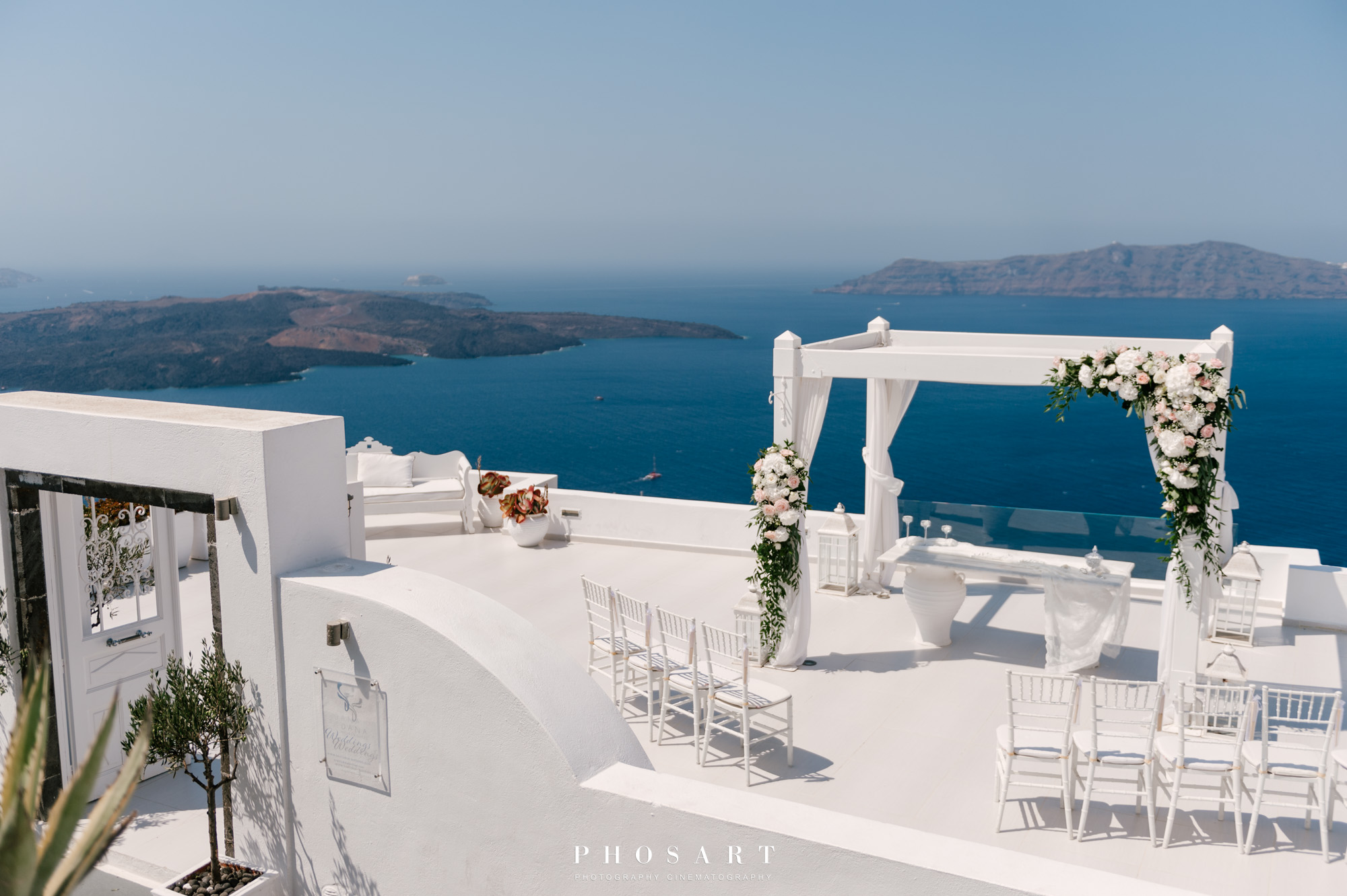 Dana Villas' large terrace above the caldera overlooking Aegean & Thirasia decorated with white furniture for a wedding ceremony