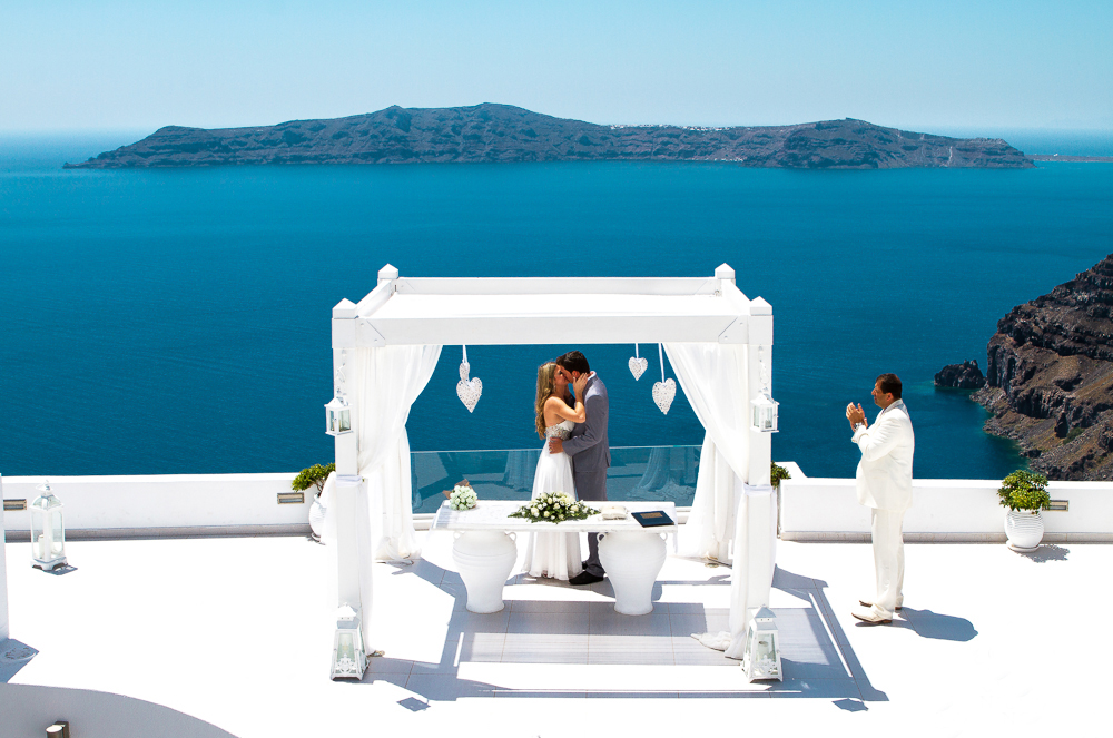 A couple gets married under a white wooden kiosk in outdoor area of ​​Dana Villas overlooking the caldera and the Aegean Sea