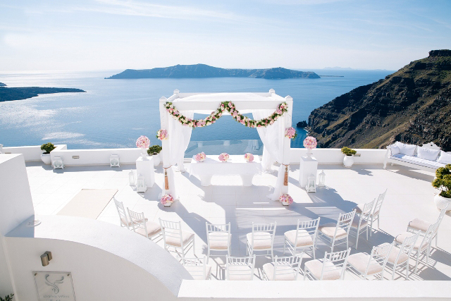 A white wooden wedding gazebo on the terrace of Dana Villas overlooking the Aegean, decorated with pink flowers